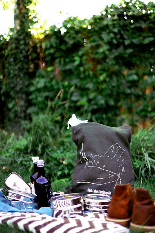 The Wasatch Tote on a picnic blanket, in a garden. There are hiking boots, and shiny metal containers carrying food items, and bottles.
