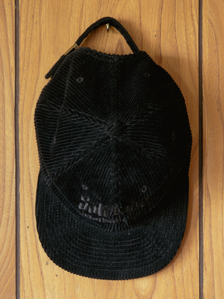 The Good Company Cap Black Corduroy hanging up on a rugged vintage wooden wall.