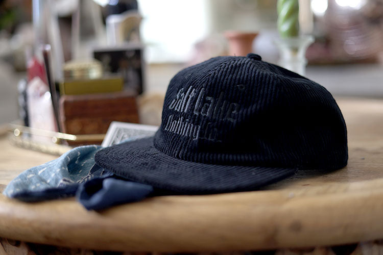 A closeup of Good Company Cap Black Corduroy, on a table surrounded by fabrics, playing cards, candles, and other ephemera.