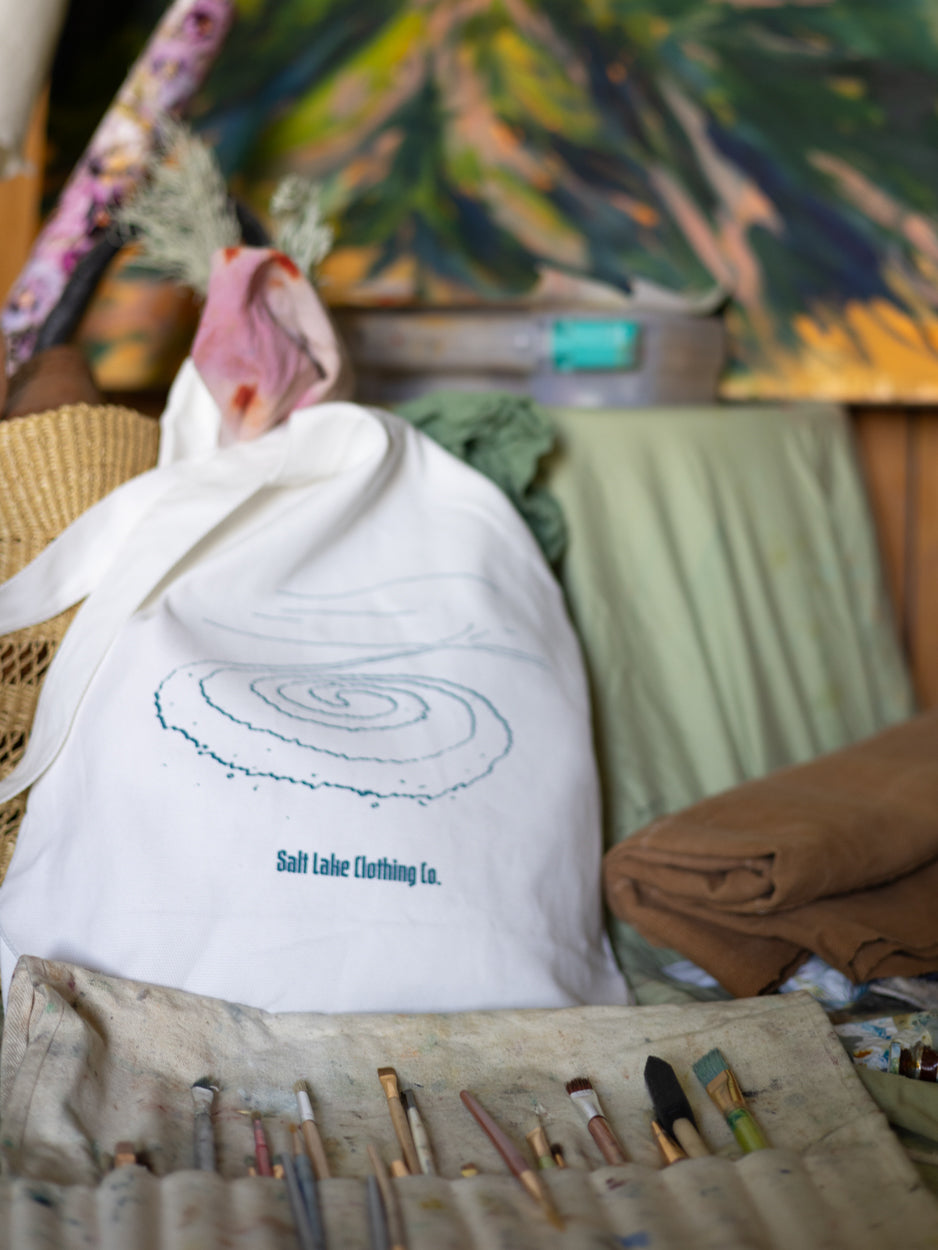Close-up of Salt Lake Clothing Jetty Tote. It is sitting in front a roll of paint brushes, in front of a still-life painting of plants.