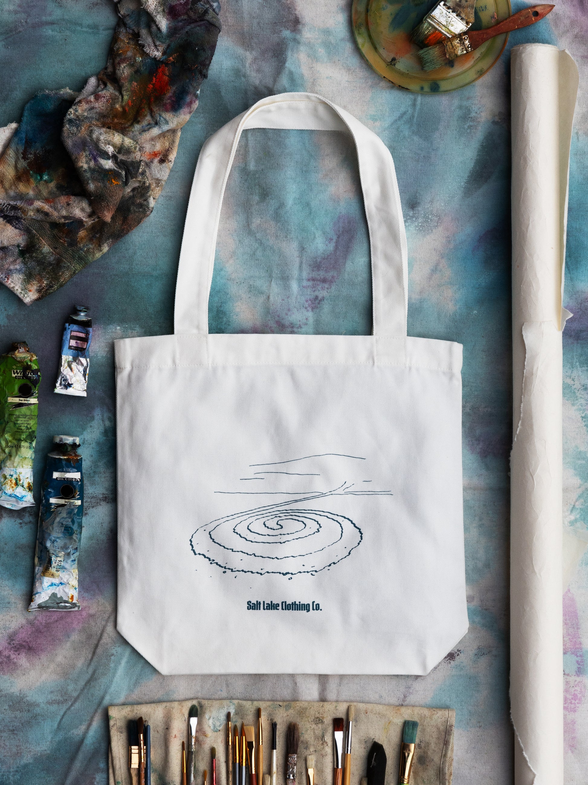Close-up of Salt Lake Clothing Jetty Tote. It is against a soft lake blue background, surrounded by paint brushes, oil paints, rags, and paper.