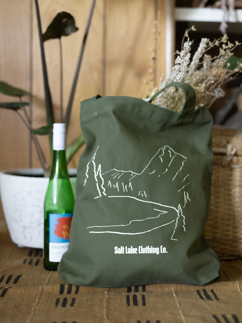 A closeup of the Salt Lake Clothing Company Wasatch Tote. It is carrying dried flowers and picnic supplies. It is surrounded by bottles for a picnic, on a picnic blanket.