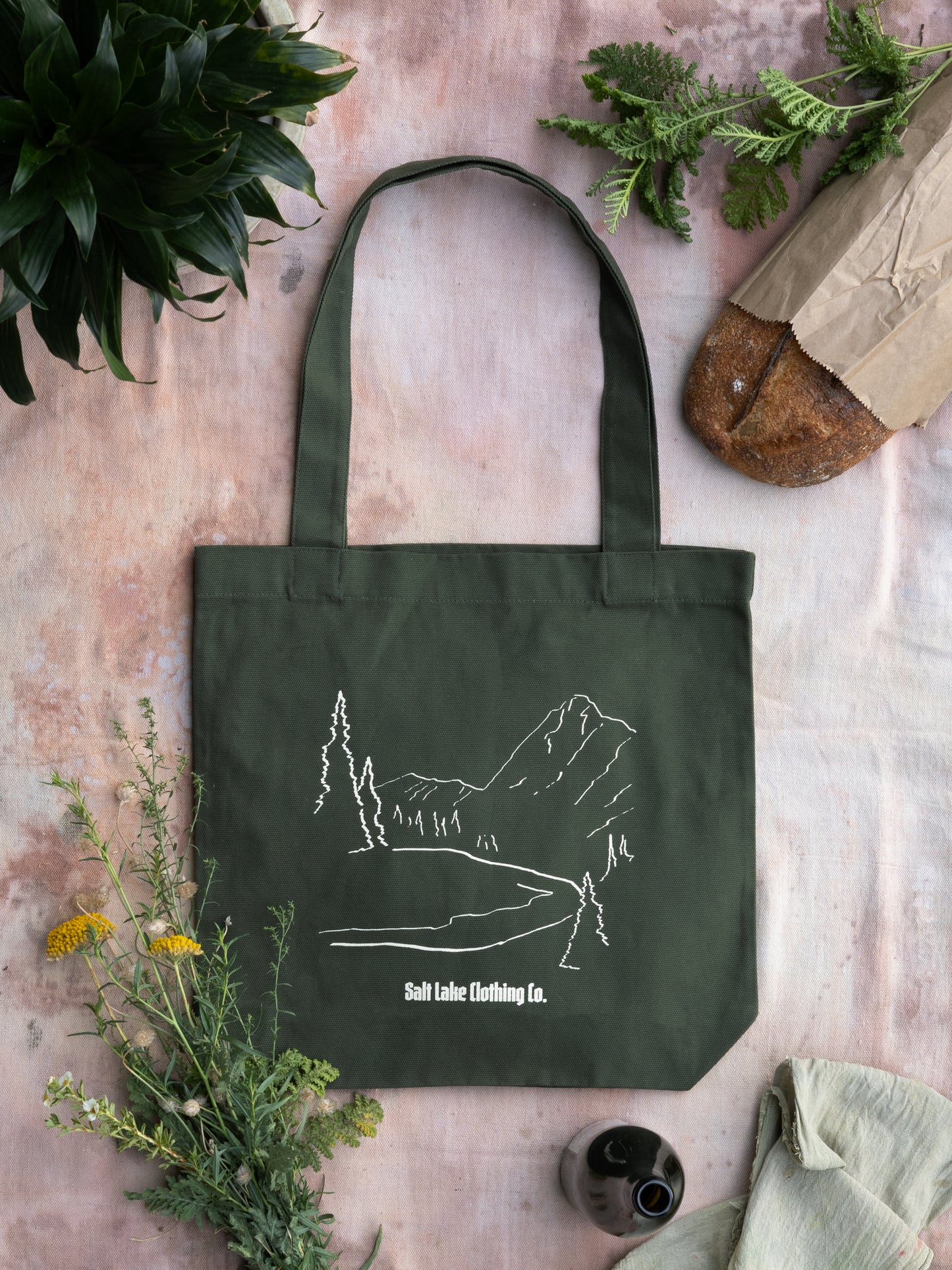 A closeup of the Wasatch Tote. It is on a soft pink drop cloth, surrounded by purchases at a farmers market: break, flowers, drinks, herbs, and a plant.