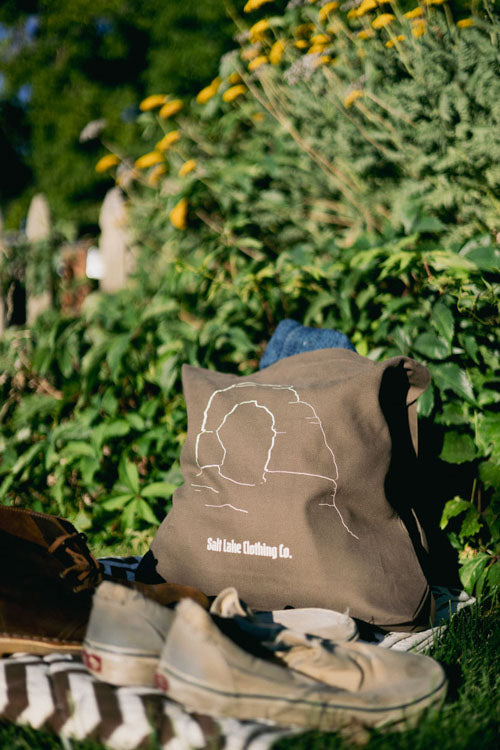 The Desert Tote sitting in a garden, on a picnic blanket, with well worn shoes. It appears to be used to carry supplies for a picnic during sunset hours.