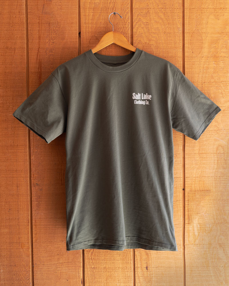 The Good Company Pocket Logo T on a hanger, against a rugged, rough hewn wooden wall.